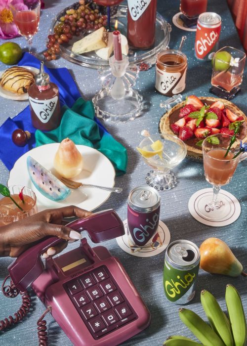 Overhead shot of a table laid out with a telephone, Ghia beverages, fruit, and snacks, styled in vibrant colors and retro fashion.