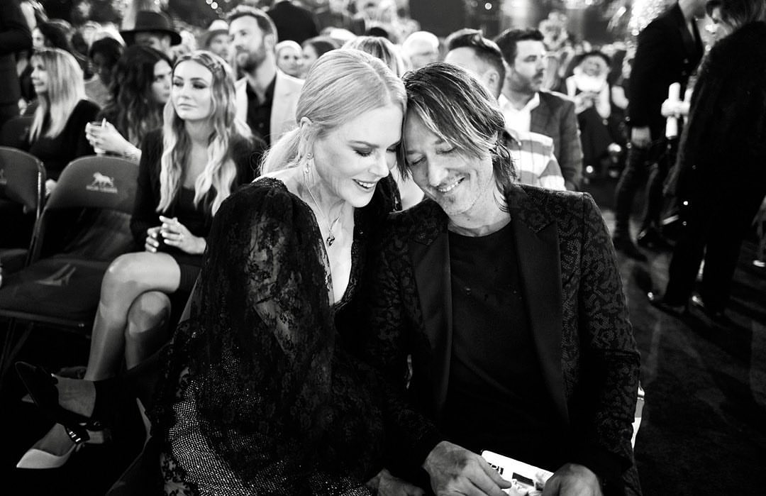 Nicole Kidman and Keith Urban attend an awards show, both seated and smiling, with a crowd of people visible in the background.