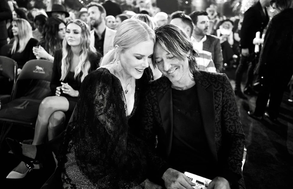 Nicole Kidman and Keith Urban attend an awards show, both seated and smiling, with a crowd of people visible in the background.