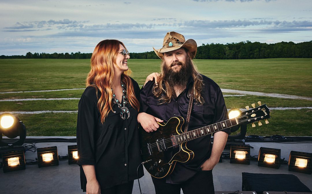 Two musicians, a woman with red hair and a bearded man in a cowboy hat, stand on a stage with an electric guitar, backed by spotlights and a field.