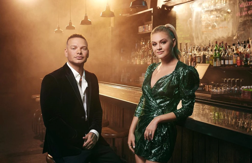 Two musicians, an adult male and adult female, pose in front of a bar with a collection of liquor bottles and decorative lighting.