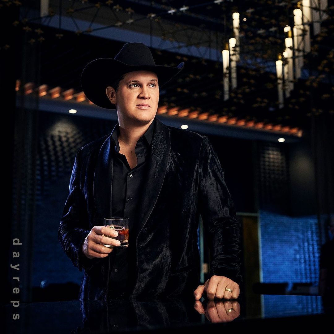 Jon Pardi poses indoors holding a drink, wearing a black cowboy hat and black velvet jacket against a blurred, softly lit background.