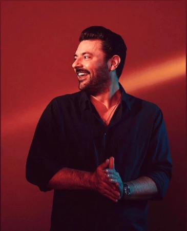 Portrait of a man in a dark button-down shirt smiling against a warm reddish background