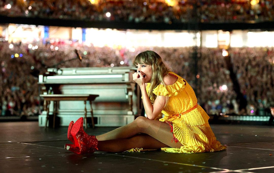 Taylor Swift sits on stage in a yellow dress and red boots, performing in front of a large crowd during a concert with piano nearby.