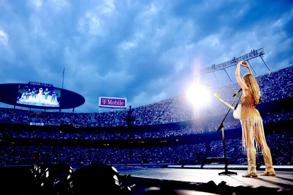Blond female guitar player performs on stage at stadium concert, wearing gold fringe outfit, arms raised with heart hand gesture to crowd.