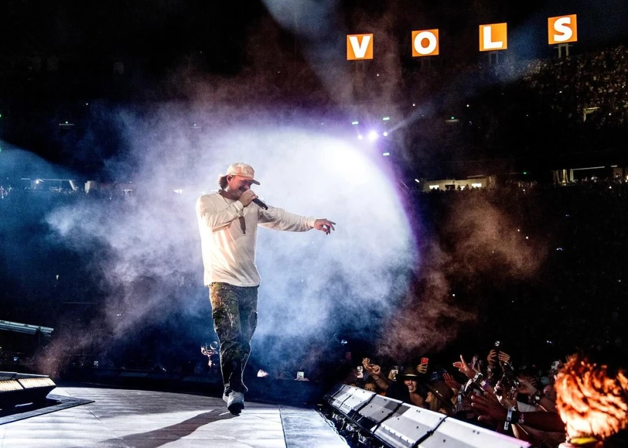 Singer performing on stage with crowd in the background. Concert features stage lighting, fog, and audience recording performance on phones.