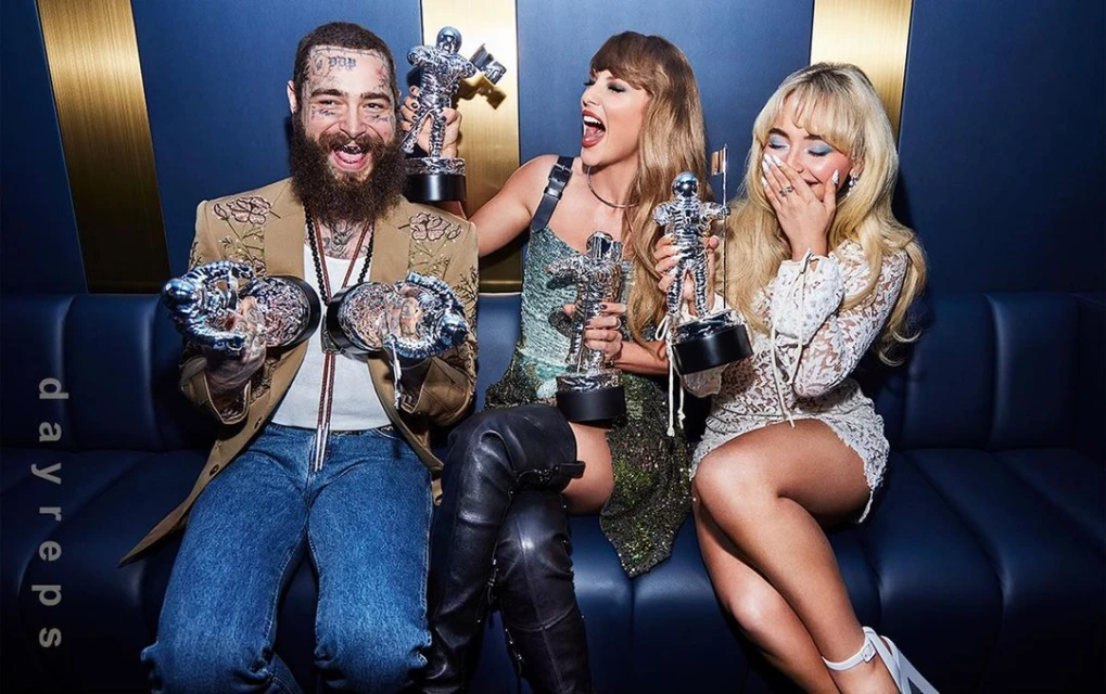 Post Malone, Taylor Swift, and Sabrina Carpenter seated on a blue couch, holding MTV Video Music Awards Moonman trophies.