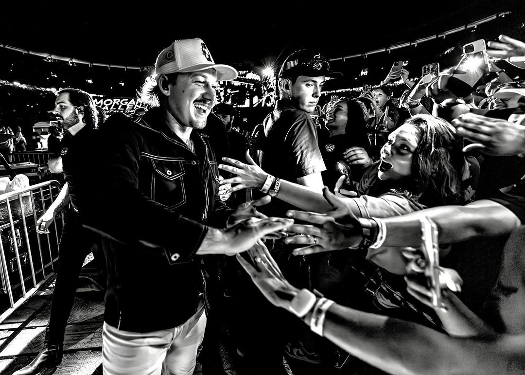 Monochrome shot of Morgan Wallen, smiling and interacting with fans, with a blurred concert crowd and stage lights in the background.