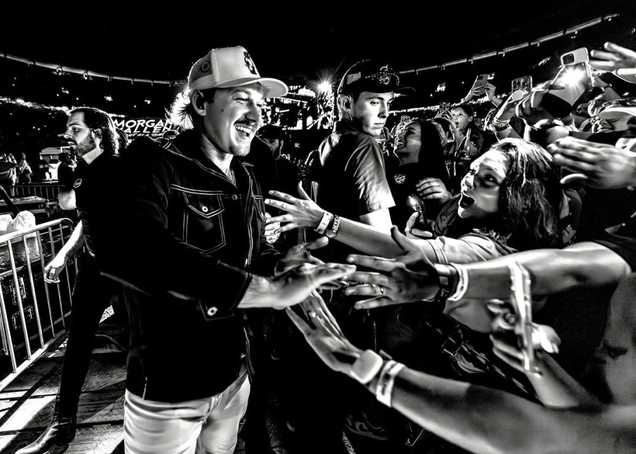 Monochrome shot of Morgan Wallen, smiling and interacting with fans, with a blurred concert crowd and stage lights in the background.