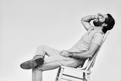 Bearded man in t-shirt and jeans laughing in a wooden chair by John Shearer