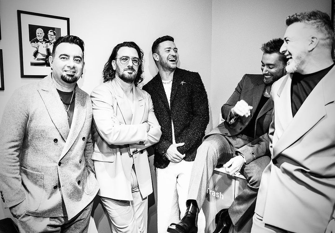 Group of five stylishly dressed men in suits posing indoors with smiles and laughter, captured in a black-and-white portrait.