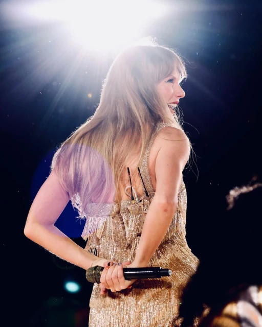 Back view of Taylor Swift on stage in gold fringe dress holding a microphone, illuminated by bright stage lights for a music performance.