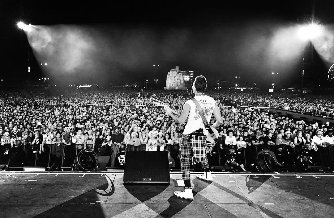 A concert featuring a performer on stage viewed from the back, facing a large crowd illuminated by stage lights in black and white.