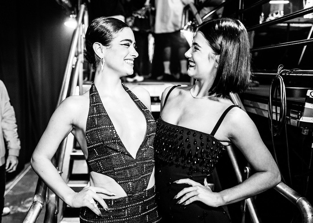 Two smiling women in dresses stand indoors, looking at each other, with visible metal rails and out-of-focus people in the background.