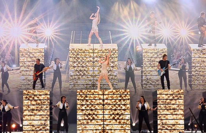 A band performs on stage with bright lights and a large audience, featuring female vocalists in sparkly dresses and musicians with guitars.
