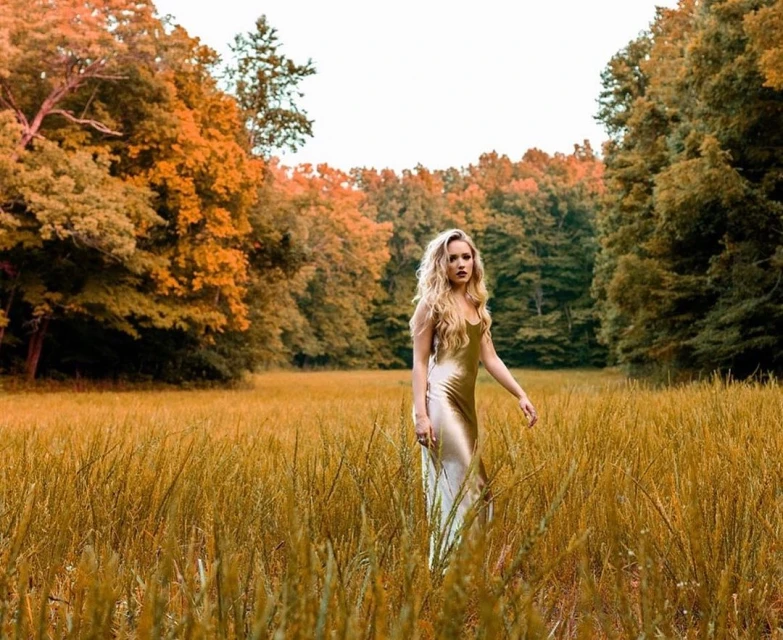 Woman with long blonde hair in a champagne-colored dress stands in a golden wheat field with a forest in the background.