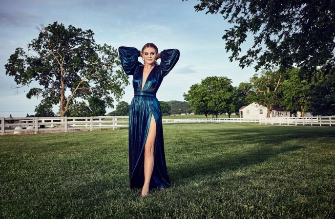 Woman in a glossy dark teal metallic gown with puffed sleeves posing in a lush green grassy field
