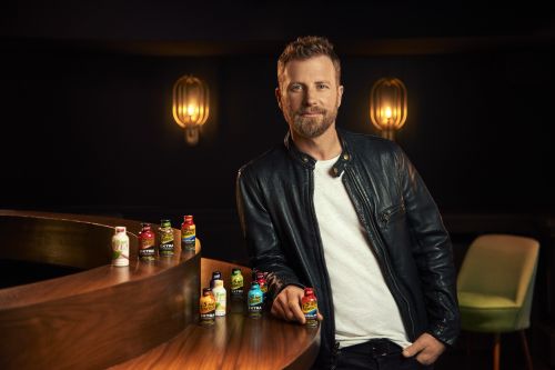 Man in a leather jacket poses with a display of energy drink bottles in a dimly lit lounge with a vintage-style decor and furniture.