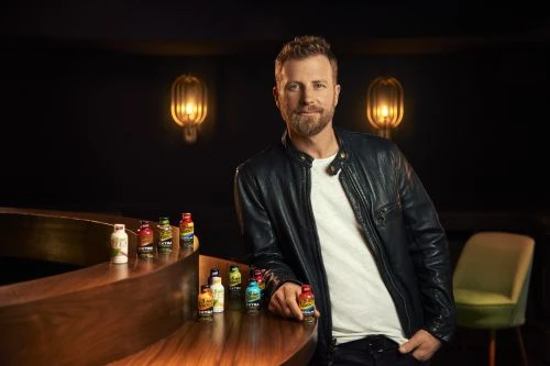 Man in a leather jacket poses with a display of energy drink bottles in a dimly lit lounge with a vintage-style decor and furniture.
