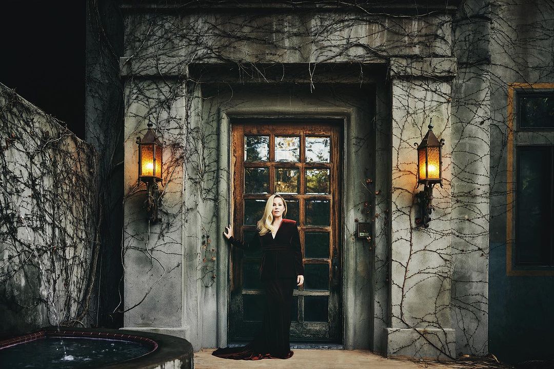 Woman in a red velvet dress stands in front of a stone building with ivy, illuminated by lanterns, evoking a mysterious atmosphere.