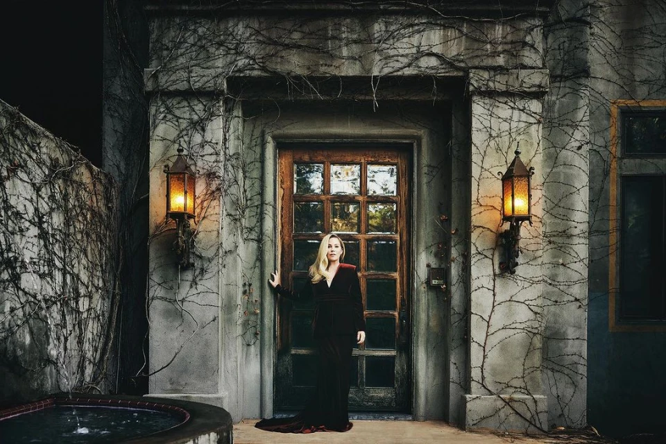 Woman in a red velvet dress stands in front of a stone building with ivy, illuminated by lanterns, evoking a mysterious atmosphere.