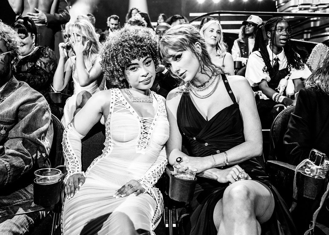 Black and white image featuring Ice Spice and Taylor Swift seated together at an event with other people in the background.