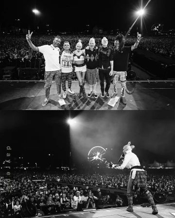 Black and white photo of a band posing on stage with arms raised, featuring a large crowd and stage lighting.