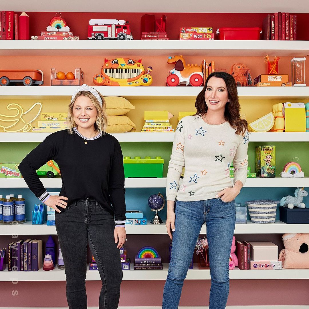 Two smiling women stand in front of a colorful bookcase filled with toys, books, and decorative objects in a cheerful indoor setting.