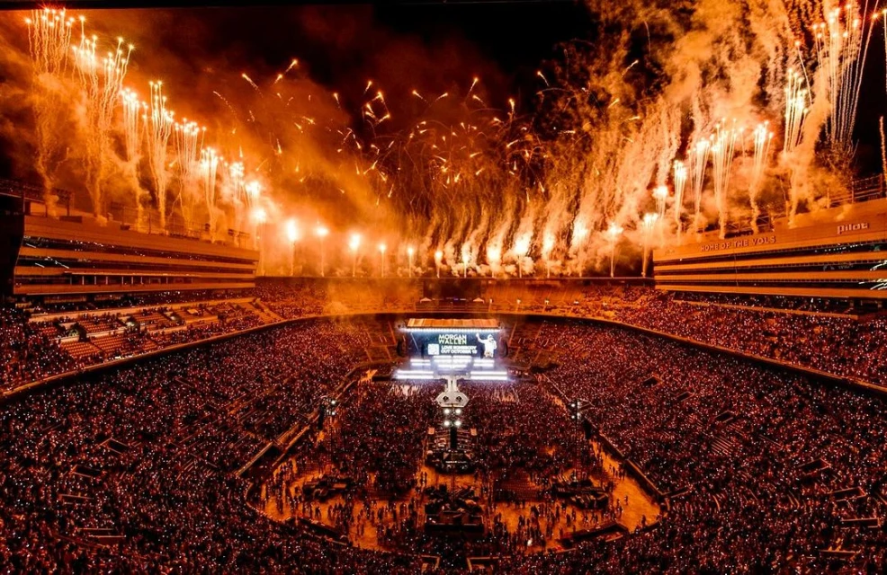 Crowd of people at a stadium event at night with an orange-tinted sky filled with fireworks above, captured in a wide outdoor shot.