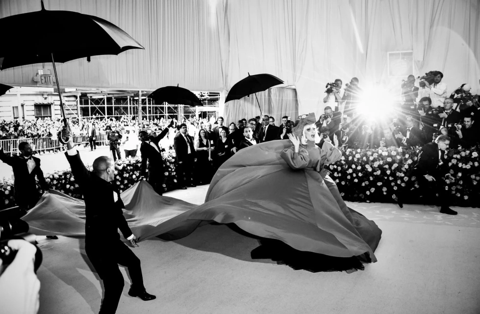 Black and white photo of a celebrity wearing a long red gown, posing on a carpet, surrounded by photographers and people with umbrellas.