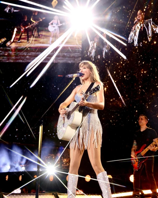 Taylor Swift performs on stage with a guitar, wearing a sparkly dress and boots, with other musicians and stage lighting in the background.