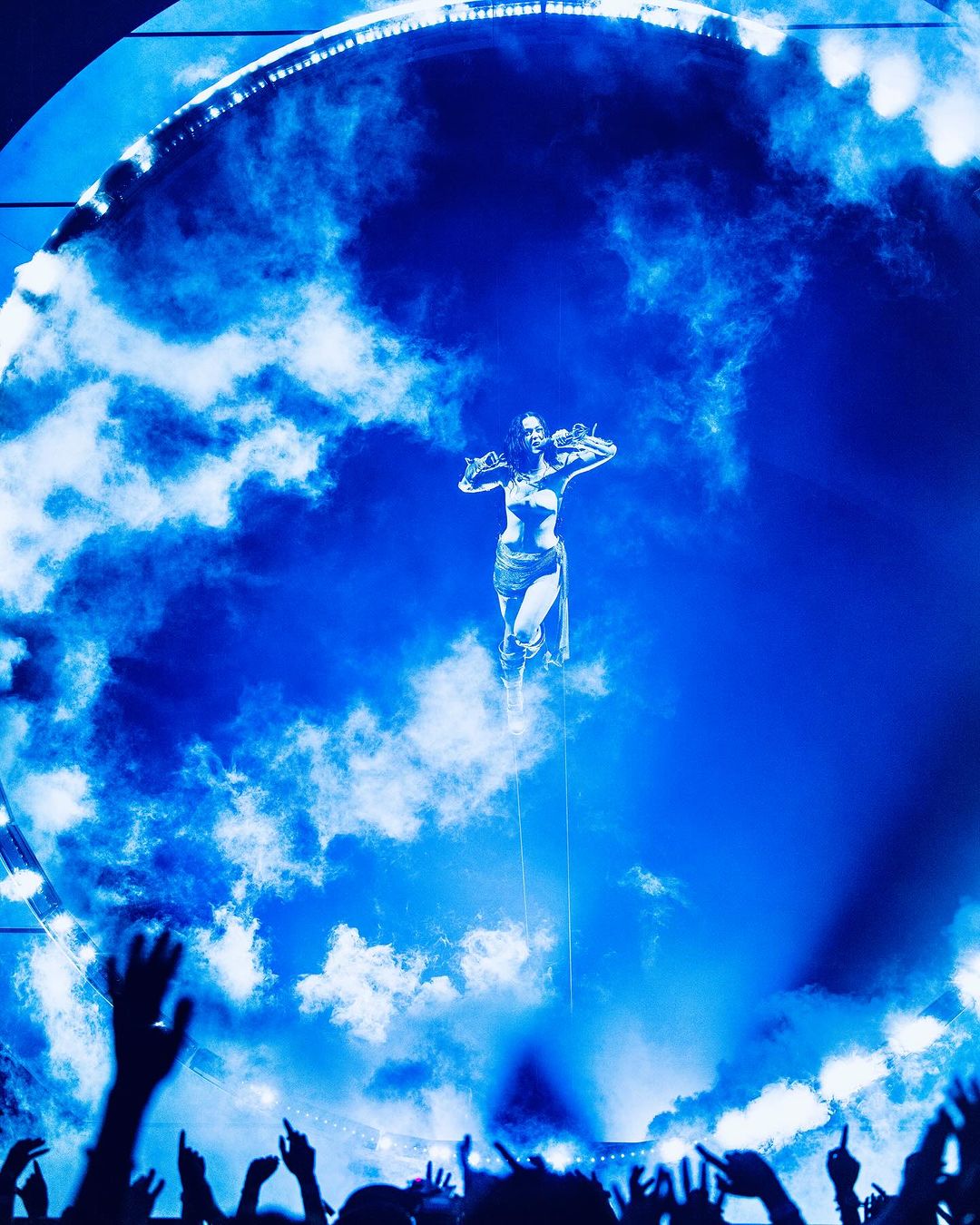 Adult female performer floats in the air on stage in front of a blue cloud-like background, with hands raised in front of silhouetted audience.