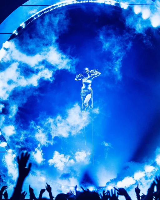 Adult female performer floats in the air on stage in front of a blue cloud-like background, with hands raised in front of silhouetted audience.