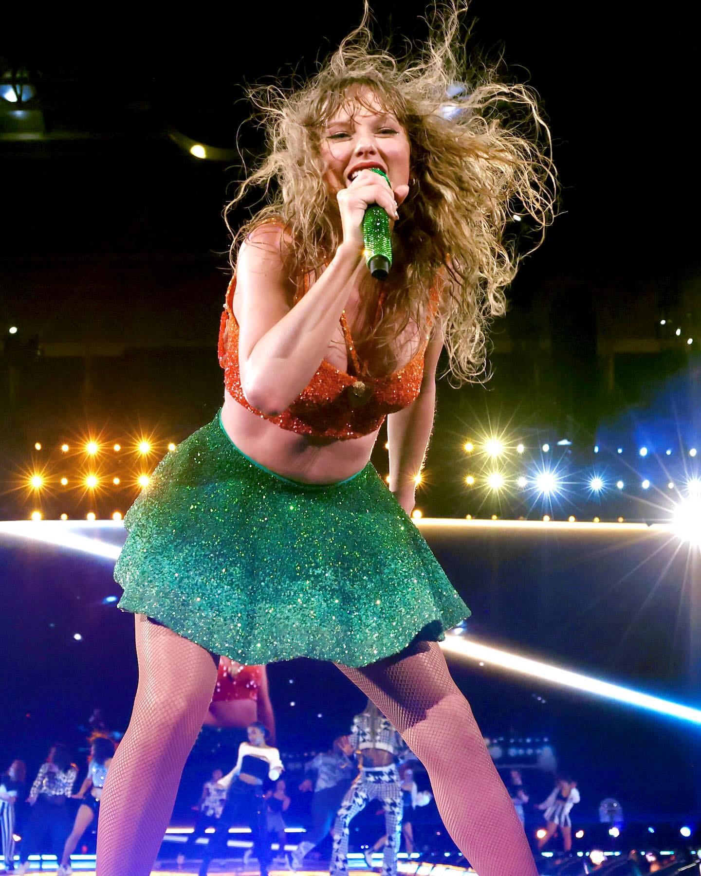 Singer Taylor Swift performs on stage with a microphone in a sparkly two-piece outfit in front of a concert crowd.