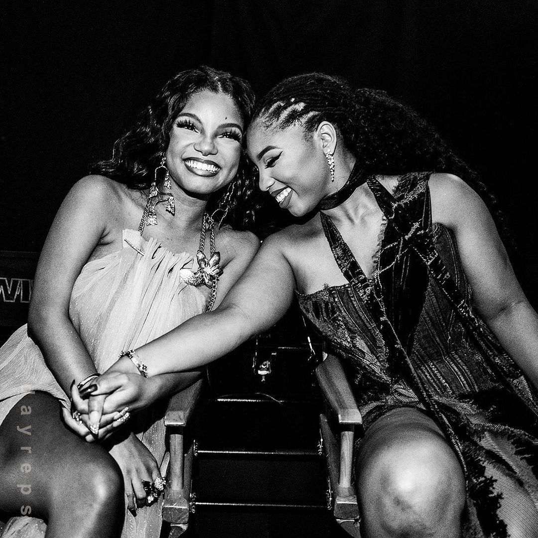 Two adult women pose sitting side-by-side holding hands, smiling in a medium indoor shot in a monochrome photo with soft lighting.