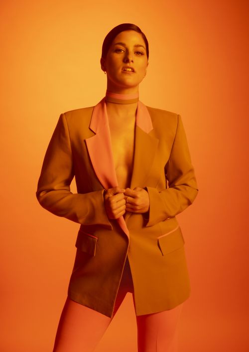 Young woman poses in a gold blazer and pink pants against a warm orange background in a fashion portrait.