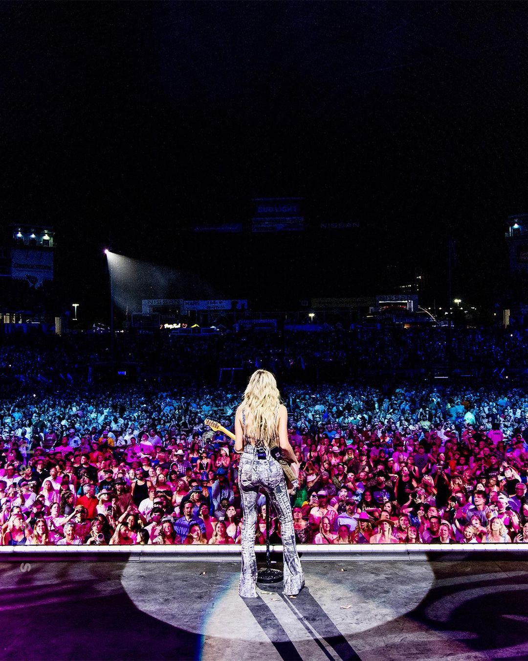 A female guitarist performs on a brightly lit stage in front of a massive audience, with an overall vibrant and energetic atmosphere.