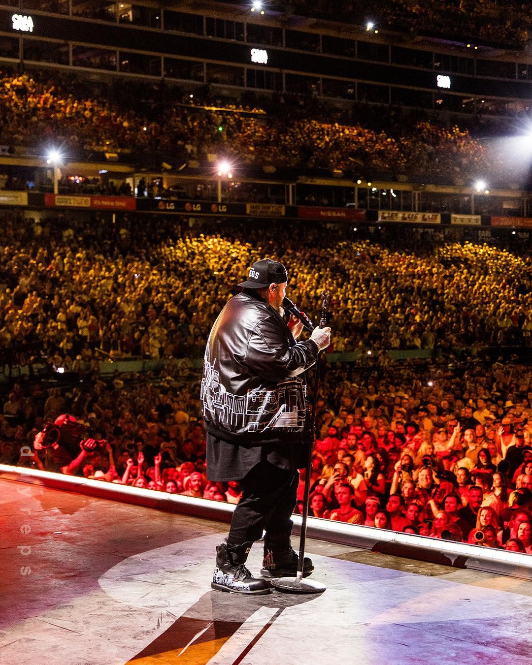 A singer performs on a brightly lit stage with a microphone, with a large crowd of people in the background at a stadium.