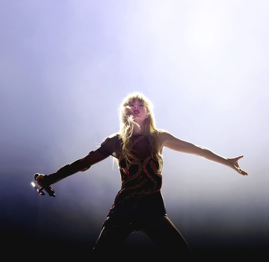 Singer Taylor Swift performing on stage with arms outstretched, wearing a dark sequined outfit against a brightly lit background.