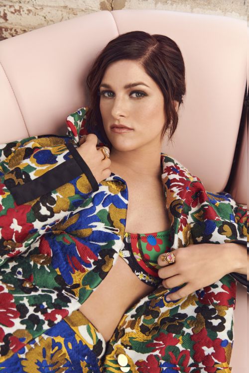 Portrait of a stylish brunette adult woman reclining on a pink couch, wearing a colorful floral jacket and bra top in an indoor setting.