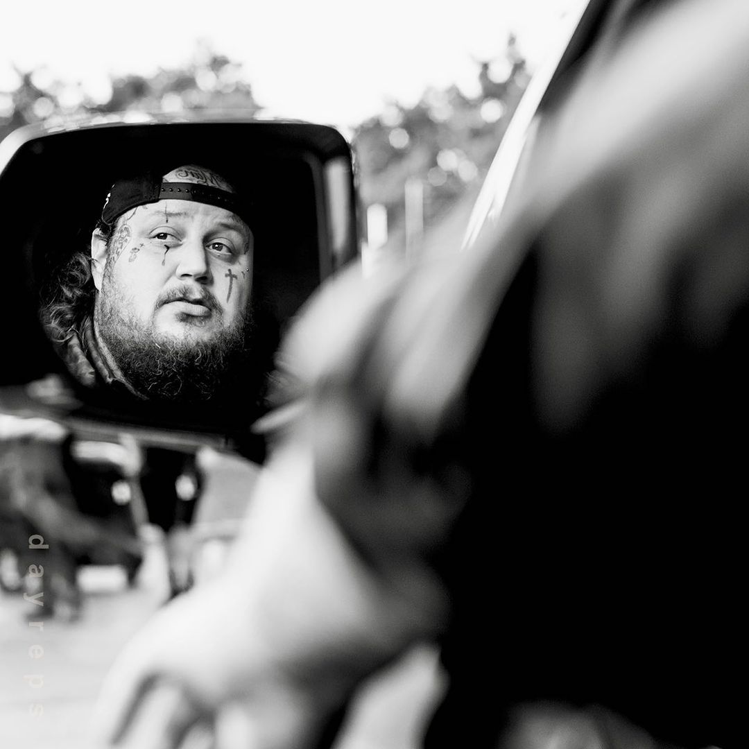 Monochrome portrait of a tattooed man in a baseball cap looking in a vehicle mirror with greenery in the background on a sunny day.