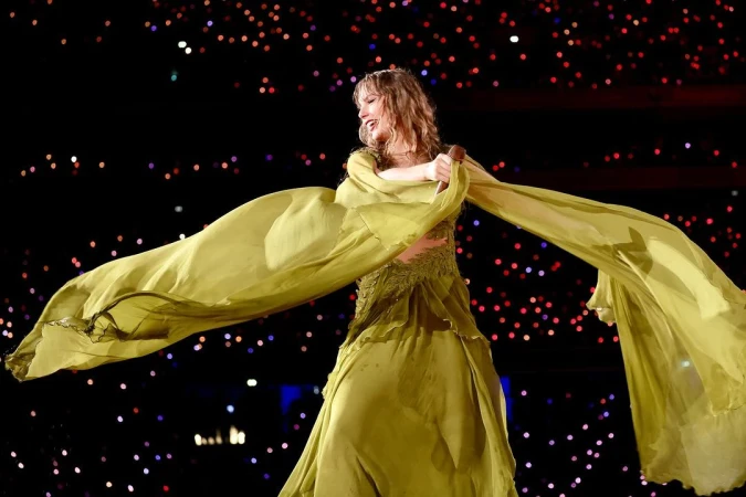 Singer Taylor Swift performs on stage in a long green dress with outstretched arms, against a dark backdrop with bokeh lights.