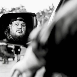 Black and white portrait of a bearded man with facial tattoos reflected in a car side mirror.