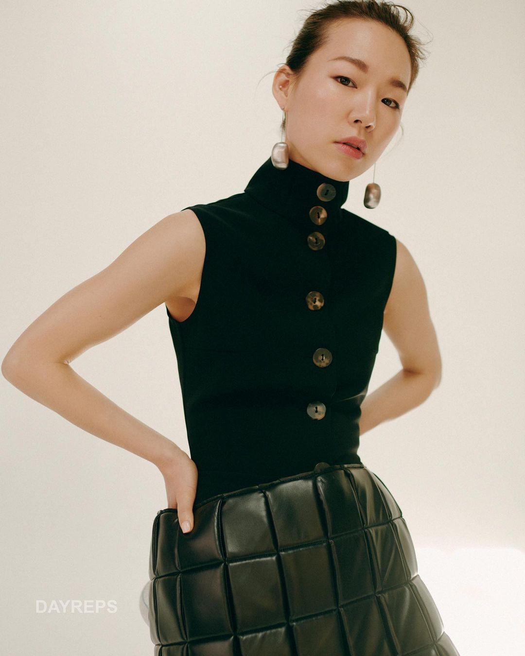 Asian woman poses wearing a sleeveless, high-neck black knit top with button detail and a black quilted, textured skirt in a studio.