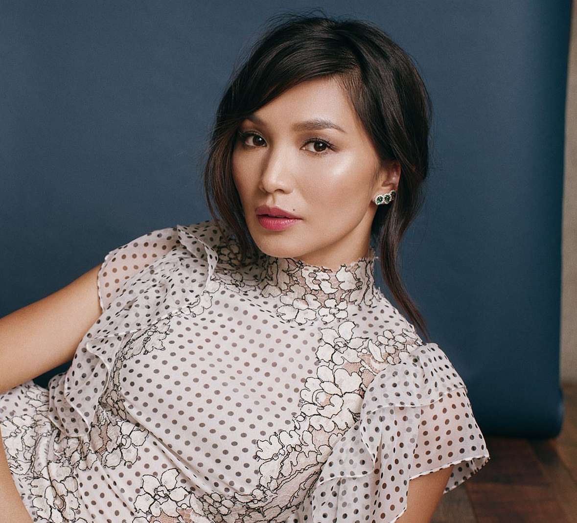 Glamorous portrait of actress Gemma Chan in a white polka dot dress against a blue backdrop, styled with dark hair and emerald earrings.