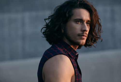 Portrait of a man with long dark hair wearing a sleeveless plaid shirt, looking off to the side against a muted grey background.