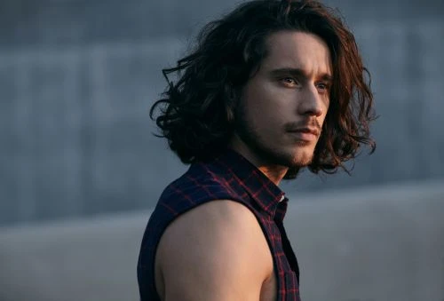Portrait of a man with long dark hair wearing a sleeveless plaid shirt, looking off to the side against a muted grey background.