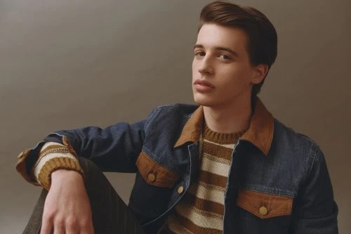 Portrait of a young adult man with styled dark hair wearing a striped sweater and a denim jacket in front of a neutral background.