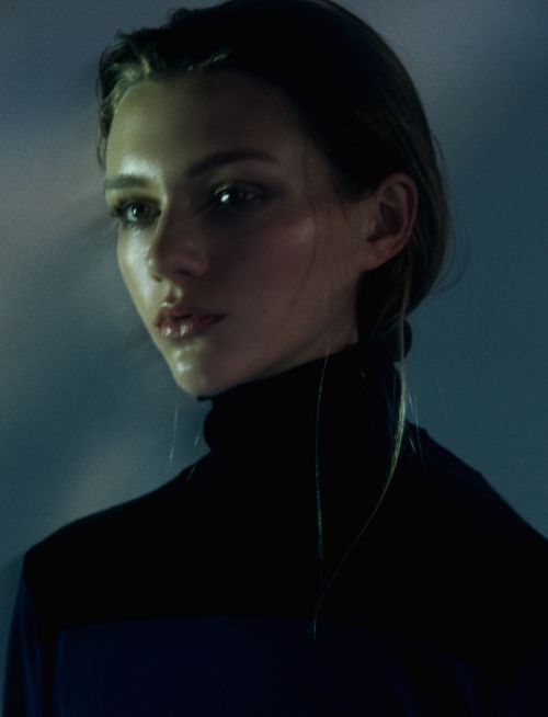 Portrait of a young adult woman with dark hair wearing a black turtleneck sweater in cool, diffused lighting against a dark background.
