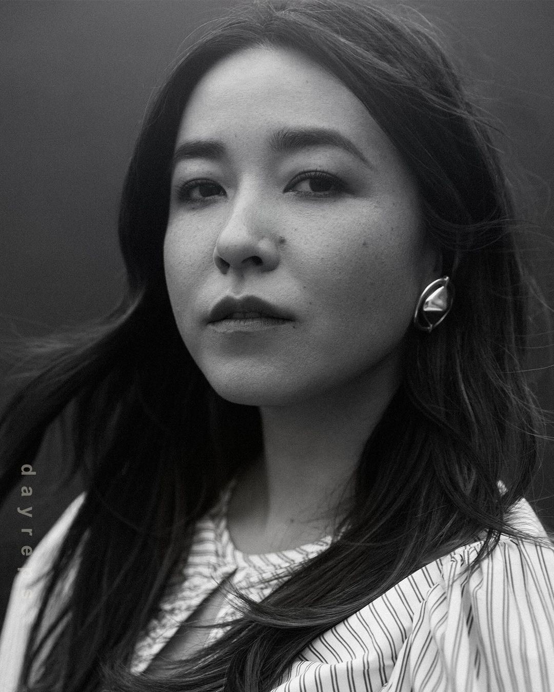 Monochrome portrait of an adult woman with long dark hair, wearing a vertically striped shirt and large geometric earring, against a dark background.